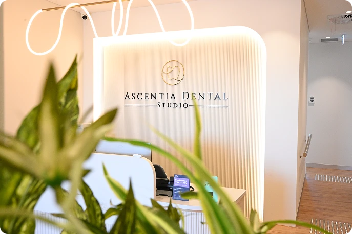 Modern dental office interior with a sign reading "Ascentia Dental Studio" on a lighted wall. Foreground features green plants, creating a welcoming atmosphere.