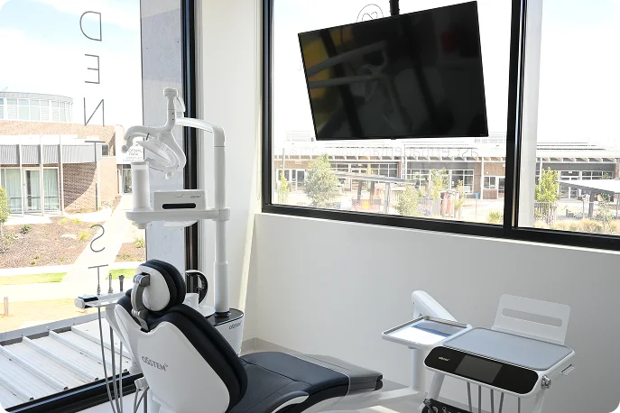 Modern dental office with a sleek chair, overhead light, and a large wall-mounted screen. Sunlit window provides a view of nearby buildings. Clean and professional ambiance.