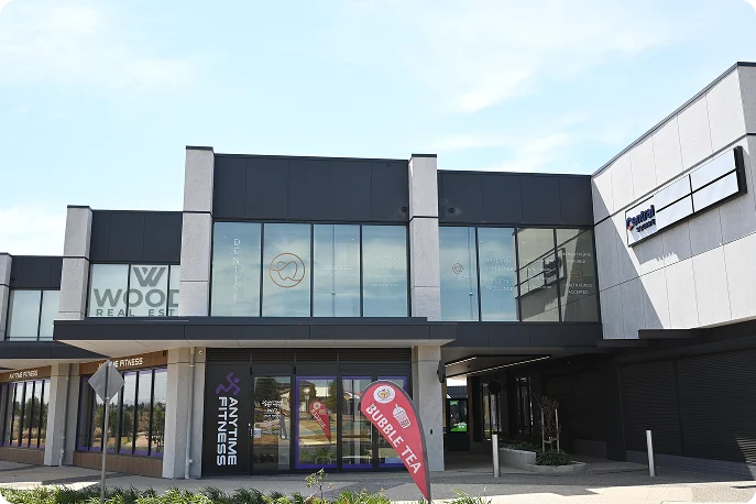 Modern commercial building with large windows, housing Anytime Fitness and a bubble tea shop with a red banner. Bright, clear sky above.
