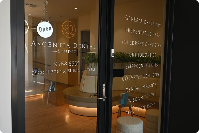 Glass door of Ascentia Dental Studio displaying services like orthodontics and cosmetic dentistry. Inside, modern, inviting decor with soft seating.