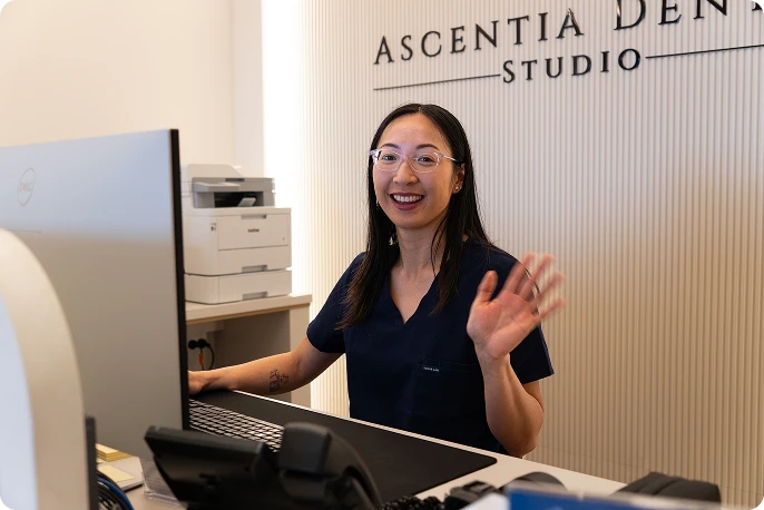 Smiling female dental receptionist at Ascentia Dental Studio, waving hello behind a clinic reception desk with computer