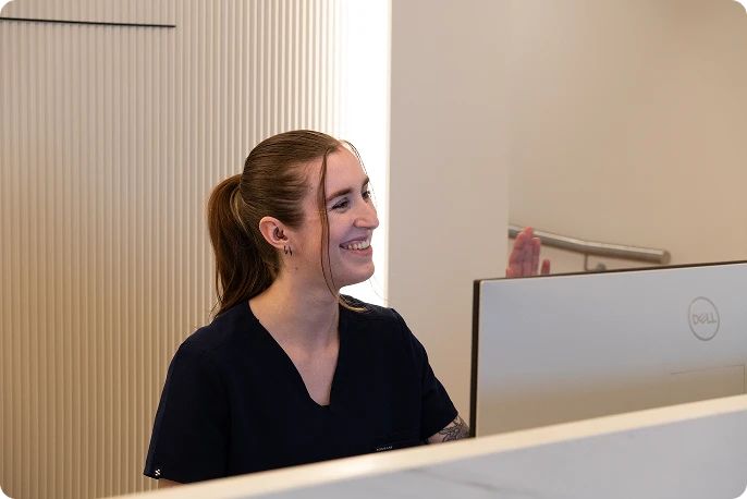 Smiling woman sits at a desk with a computer, warmly engaging with someone off-frame. 