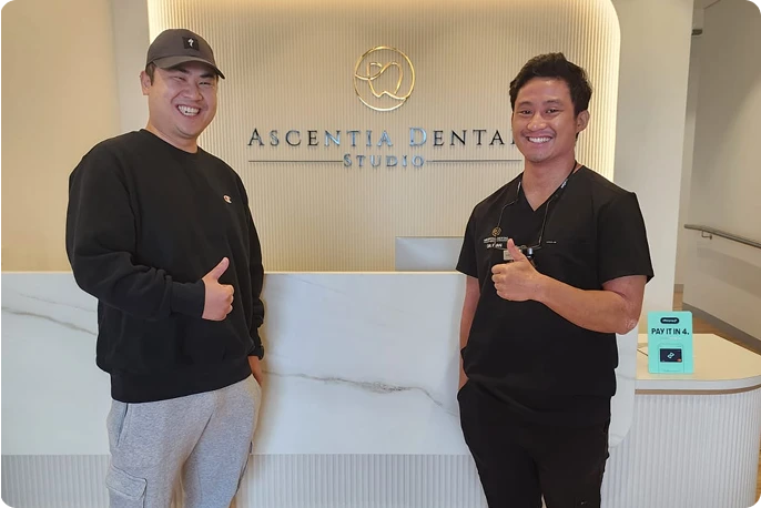Two smiling men stand in front of a reception desk at Ascentia Dental Studio, both giving thumbs up. 