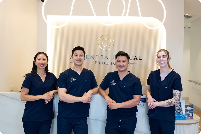 A group of four dental professionals in navy scrubs stand smiling at a reception desk.
